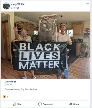 Two men pose with a black lives matter banner. The man on the right holds up a large photo of Hitler and the man on the left gives a seig heil. They're standing inside a house's kitchen.