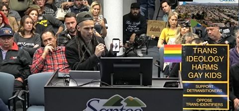 This is a screenshot of a livestream of a Santee City Council meeting. In it, Shaun Frederickson, an antivaxx organizer and Awaken Church affiliate, addresses city council. He is a white man with dreadlocks. Shaun is holding his phone up to the council, and in it one can barely see the figure of a woman. This is a video of a trans sex-worker's adult content from twitter. Behind Shaun, there are numerous attendees, including Chris Reyes, who is squatting with a camera. To Shaun's left—our right—is a transphobic sign. It reads: "TRANS IDEOLOGY HARMS GAY KIDS. SUPPORT DETRANSITIONERS. OPPOSE TRANS-IDENTIFIERS."