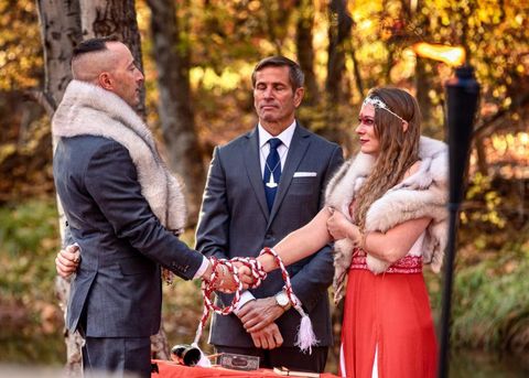 Ryan Drago wearing a gray suit and an animal pelt while holding the hand of Ashley Krogstad, who is wearing a red and white lace dress with the same style of animal pelt as Drago. Her eyes are painted red and she's wearing a white tiara. Their hands are linked with a white and red braided rope. In between them is a table with a horn and hammer and Joseph Rozanek wearing a gray suit and large Thor's Hammer pendant. They are surrounded by forest scenery.