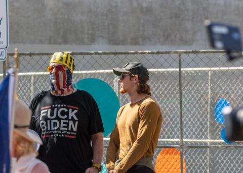 The Proud Boy on the left is wearing a black shirt that reads, "fuck Biden." He has an American Flag gaiter covering his face, and a black and yellow gaiter covering the top of his head.