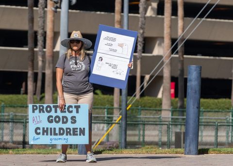 A protester stands on the corner opposite the New Children's Museum. They have two signs. One sign is held low, and it reads "protect all god's children from drag queens." The other sign is raised, and has a transphobic meme.