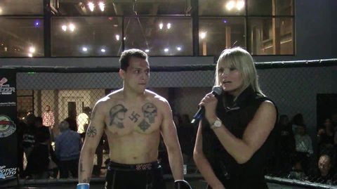 A man in a boxing ring stands with his shirt off next to a woman with a microphone. The man is shirtless with a swastika tattoo on his chest along with a celtic cross and other Nazi imagery.