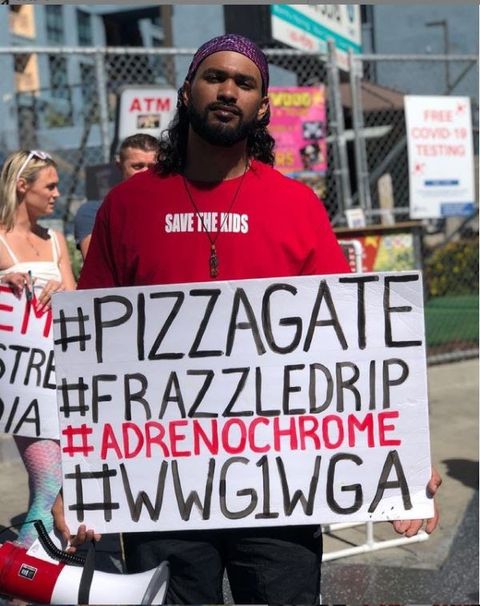 Thaddis Johnson, a bearded man in a red shirt with 'save the children' emblazoned on it, holding up a cardboard sign with the hashtags 'Pizzagate,' 'frazzle drip,' and 'WWG1WGA.'