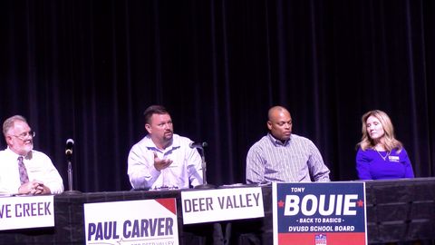  Paul Carver, wearing a white collar shirt, speaks in a microphone to an unpictured crowd. He’s holding his hand up as he speaks. Next to him are other republican candidates for school boards. They’re all sitting on a stage with black drapes behind them and black tablecloths on the tables they’re sitting in front of. Plain white signs for “Cave Creek” and “Deer Valley” school board are taped to the tables. Paul Carver has his red, white, and blue campaign sign taped in front of his table.