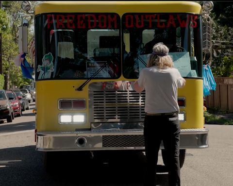 A person stands in the middle of a street, blocking the path of a fake fire truck with 'Freedom Outlaws' and 'FAFO' stenciled on the front. They are covering their ears.