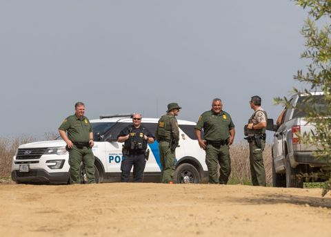  the foreground consists of a dirt road, out of focus. In the distance, three Border Patrol agents confer with a Federal Protective Service Officer and a State Park ranger. Behind them is a Federal Protective Service cruiser.