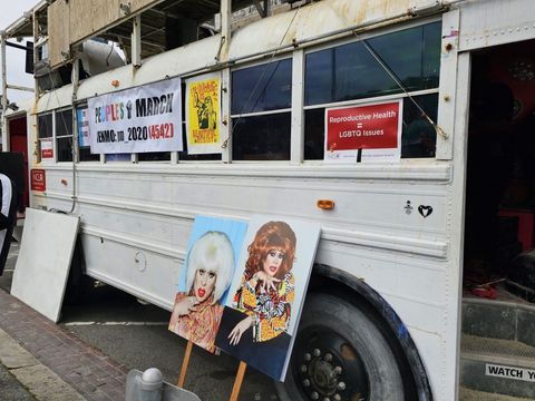 A white school bus with signs with photos of a drag queen in colorful patterns. the bus has a banner that says "people's march" and another sign that says "reproductive health equals LGBTQ issues"