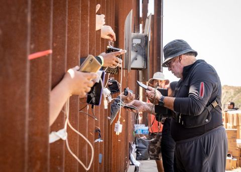 an activist plugs in a charger into peoples' cell phones as they stick their phones through the border fence
