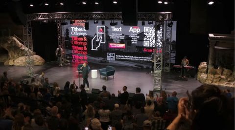 A man stands on a stage as people applaud and another man enters from behind the stage. the screen behind them has a QR code and other links to donate tithes and offerings