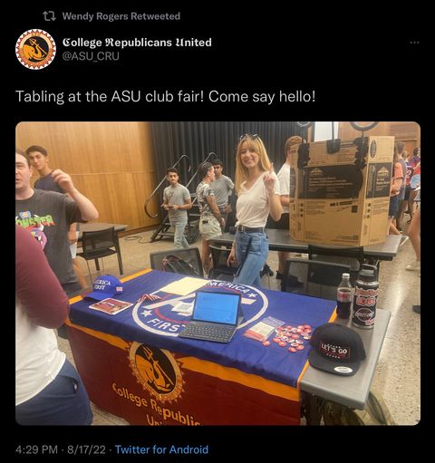 a screenshot off twitter from CRU with the caption 'tabling at the ASU club fair! Come say hello!' A photo of a woman posing with her finger in the air accompanies the post, she’s posing in front of a table with the ASU-CRU flag and the blue America First flag. Two hats also sit on the table, one is a blue America First hat, the other is a black Let’s Go Brandon hat. Dated August 17th 2022, retweeted by Wendy Rogers