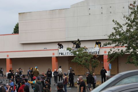A crowd gathers in a parking lot of an abandoned big box store as three men drop a banner from its roof that says 'free our political prisoners'