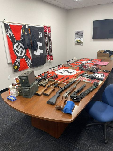 A collection of Nazi memorobilia on a table with nazi flags hanging on wall and shotguns, rifles and ammo piled on the side of the table