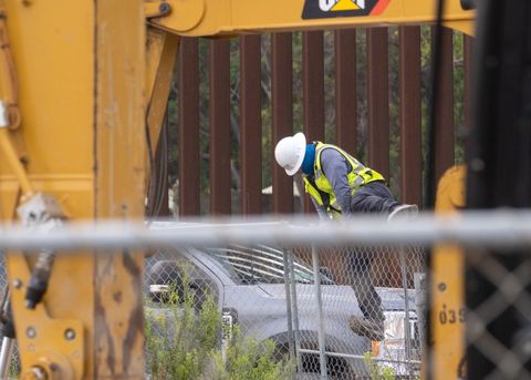 An excavator and a chain link fence border the frame in the foreground. In the middle distance, a Spencer employee in a white hard hat and green high visibility vest hops the fence. Beyond him, one can see the border wall.