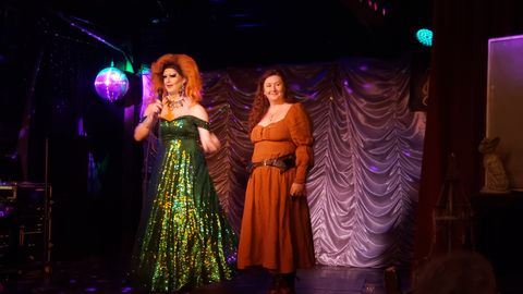 A photograph of two people on the stage inside Old Nick's Pub. On the left, a drag queen with a big orange wig wearing a sparkly green dress. On the right, Emily Chappell with her brown hair and wearing a pumpkin orange dress with brown belt. Bitchcock, the drag queen, is speaking into a microphone.