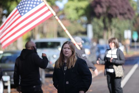 Audra Price, with her medium long dirty blonde hair, wears a black sweatshirt. She's standing, facing the camera. Directly behind her is Richard Elce holding a large American Flag by a bamboo flagpole. To her left is the bald, but bearded, Proud Boy Barry Johnson Jr.