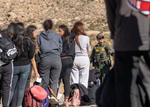 In the foreground, asylum seekers stand. They're out of focus. In the midground, more asylum seekers are standing. They have long hair, various kinds of warm clothing, and bags. Just beyond these asylum seekers stands one border patrol agent. In the background, one sees desert sand and brush.