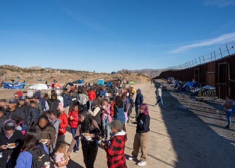 A crowd forms a wide queue ahead of the camera. There are hundreds. To the right of the frame, the southern border wall is visible. It casts a shadow and tents are pitched against it. In the distance, one can see desert hills. Numerous tents dot the detention site.