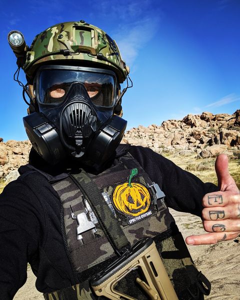 Zamudio poses in a gas mask and body armor giving a 'surf's up' hand gesture. There's tattoos on his fingers that spell out 'white boy.'