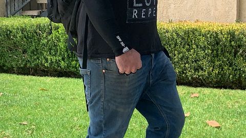a man, cropped from his upper torso to his knees, walks along a sidewalk next to a building. He's wearing jeans and a black shirt with long sleeves and his fist is clenched around brass knuckles