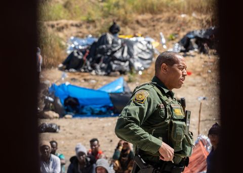 through the bars of the border wall we see a border patrol agent holds something near his utility belt and squints while talking to someone out of view. asylum seekers sit in the dirt and look at him
