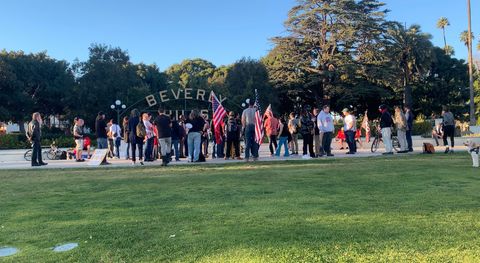 about thirty or so people gathering around a sign in a park that says 'Beverly Hills' while a dog in the foreground looks at them