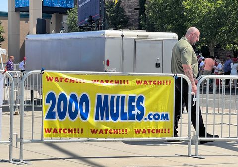  the metal line fencing displaying a simple yellow banner with large blue text reading 2000MULES.com and “WATCH!!! WATCH!!! WATCH!!!” on the top and bottom. A man is walking by, some can be seen standing around in line.
