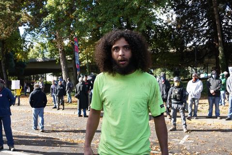 Marcus Edwards wearing a bright lime green shirt stares into the camera with a crazed look in his eyes. Behind him, a plethora of bigots and Nazis stand on the other side of the street.
