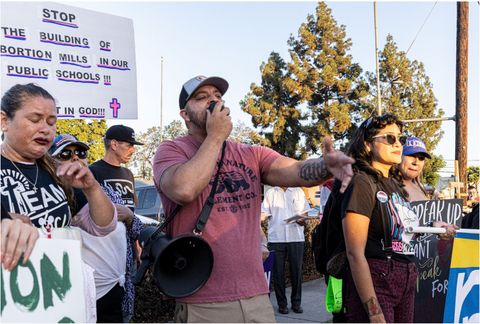 a man pointing with a bullhorn slung over his shoulder in a crowd of other people holding signs with slogans like stop the building of abortion mills in our public schools