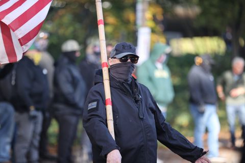 Elce wears all black and a hat, glasses, and gator covering his face. He's holding his bamboo flagpole and the American Flag at the end of it can be seen in the left corner of the shot. Blurred, behind him, are other bigots at the protest.