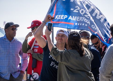 Cordie Williams poses for a video and a photo with an attendee, rhythmically swaying to the generic dance drum break being played on loop after the two speakers finished up. A trump flag flows in the breeze above his head, held by a man in a separate conversation in the background.