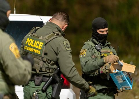 a masked border patrol agent takes an object out of a cardboard box