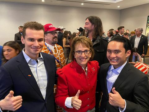 In a crowded room, state senator Wendy Rogers poses for a photo with far-right commentator Jack Francis aka Red Eagle Patriot and APU co-founder Vince Dao. They all smile and give a thumbs up.