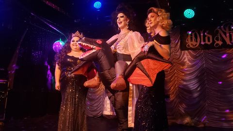 Three drag queens stand on the Old Nick's Pun stage after their drag queen story time event. Sunshine, on the left, is wearing a sparkly dark dress and gold-colored crown. Maliena, in the middle, is wearing a dress and also in a blow-up dragon costume as if she's riding it around. Elekra, on the right is wearing a sparkly black dress and has their left arm curved inward in pose.