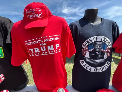 two shirts and a hat on sale being displayed at the merch booths. One shirt is bright red with white text reading 'Prescott, Arizona for Trump. Save America.' The red hat sitting on top matches the shirt in color and text. Another shirt being displayed next to the red shirt is a black spin on a Sons of Anarchy shirt with Trump photoshopped as a biker. The text reads 'Sons of Trump, MAGA Chapter.'
