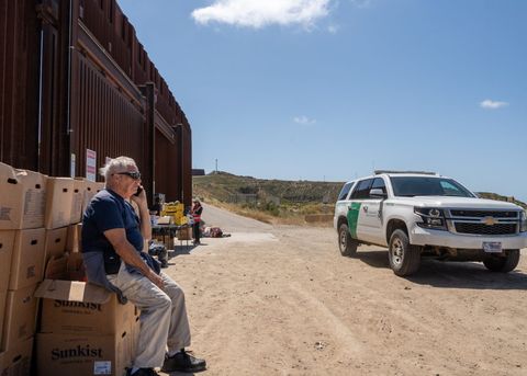 an activist sits on a box of supplies next to a border patrol car 