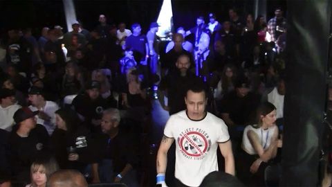A man in a white shirt with a red 'no' sign with images of drugs in the middle and the slogan "reject poison" walks through a crowd in a fight arena.