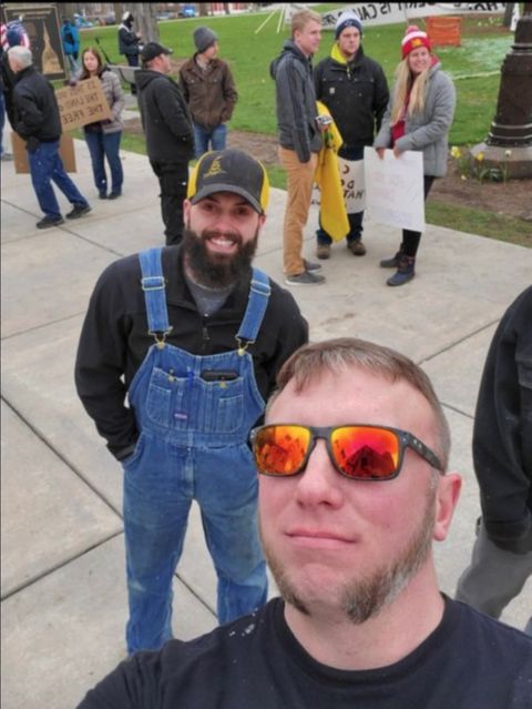 A man with sunglasses and utton chops poses next to a bearded smiling man in a yellow and black ballcap with the don't tread on me snake on it at a rally with people holding signs behind him