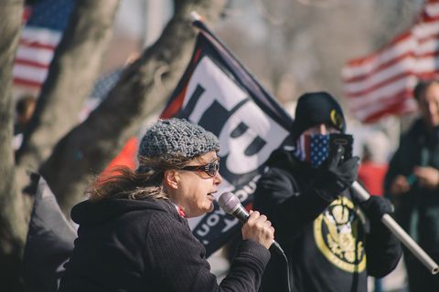 a woman at a podium talks to a crowd as a man in a yellow and black proud boys hoodie with his face hidden by a mask stands by and records her with a phone