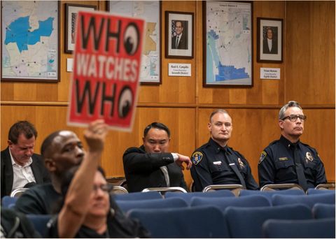 officers in the back row look on as an attendee raises a sign. The sign reads, "who watches who."
