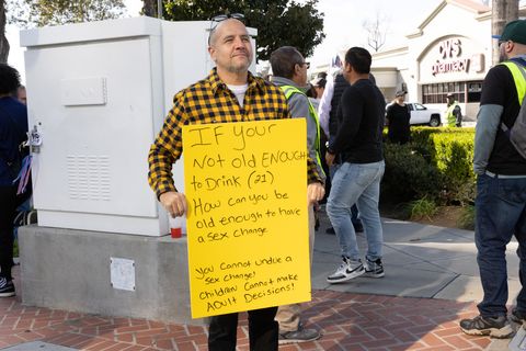 a man stands with a sign in front of a CVS. the sign says "if your not old enough to drink (21) how can you be old enough to have a sex change you cannot undue a sex change! children cannot make Adult Decisions!
