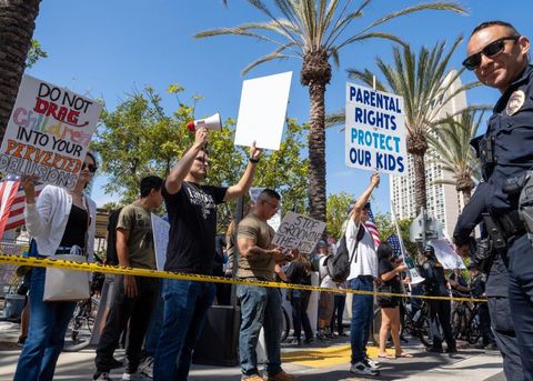 This image is shot from below, angled upward. On the right side of the frame, and officer is looking down into the camera while smirking. The rest of the shot consists of a crowd of protesters just behind caution tape, LEXIT members and a Proud Boy among them. There are palm trees and clear blue skies.