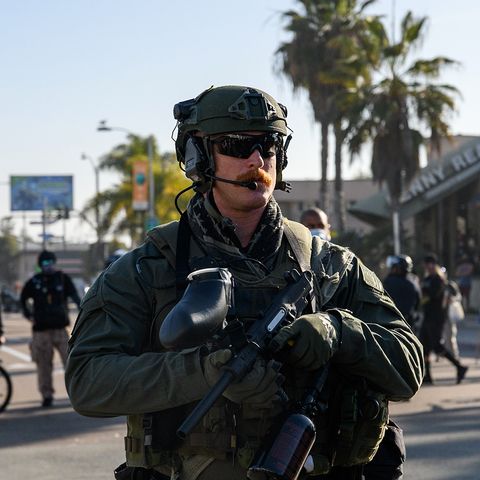 A SWAT officer stands with a pepper ball gun and a big mustache.