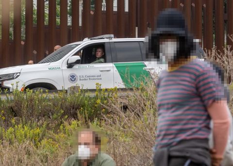 Protesters are visible in the foreground, but are out of focus. Further back, on a road above the protesters, a Border Patrol agent sits in his marked cruiser, observing.