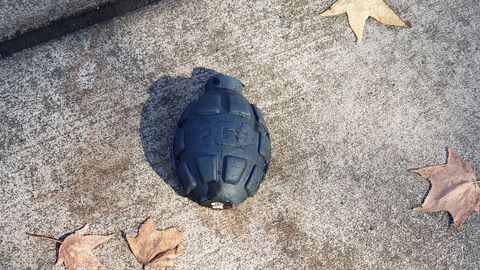 A photograph of half a plastic grenade-shape shell on the ground. It has the logo for Enola Gaye molded on the side and it's printed marking on the bottom. Around it, a handful of dry brown leaves are scattered on the ground.