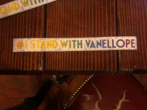 A photograph looking down at a table with two stickers laying on top. They are long strips and the font in a rainbow gradient. They read 'I STAND WITH VANELLOPE'