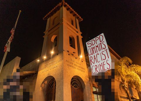 two protesters stand in front of the SDPD Central Division substation. The protesters are pixelated to protect their identity. One protester, on the right side of the image, is holding a sign. The sign is white with red text that reads 'SDPD Commits Racist Crimes.'
