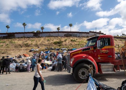 people walk around on a hill below the border fence near a semi truck