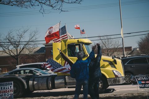 two people wave american flags at a yellow semi truck that's got a polish and american flag on each side.