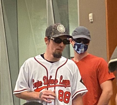 A nazi in a black sun baseball cap with a baseball jersey that says goyim defense league 88 with a masked nazi behind him