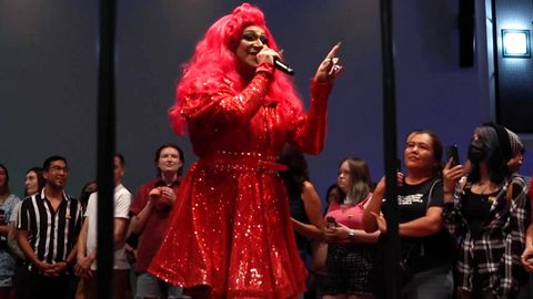 drag queen Pyraddiction is dressed in a bright red wig and bright glittery red long-sleeve dress as she smiles and holds a mic while addressing the crowd. The crowd looks on, many holding up with phones filming, and listens to Pyraddiction speak.